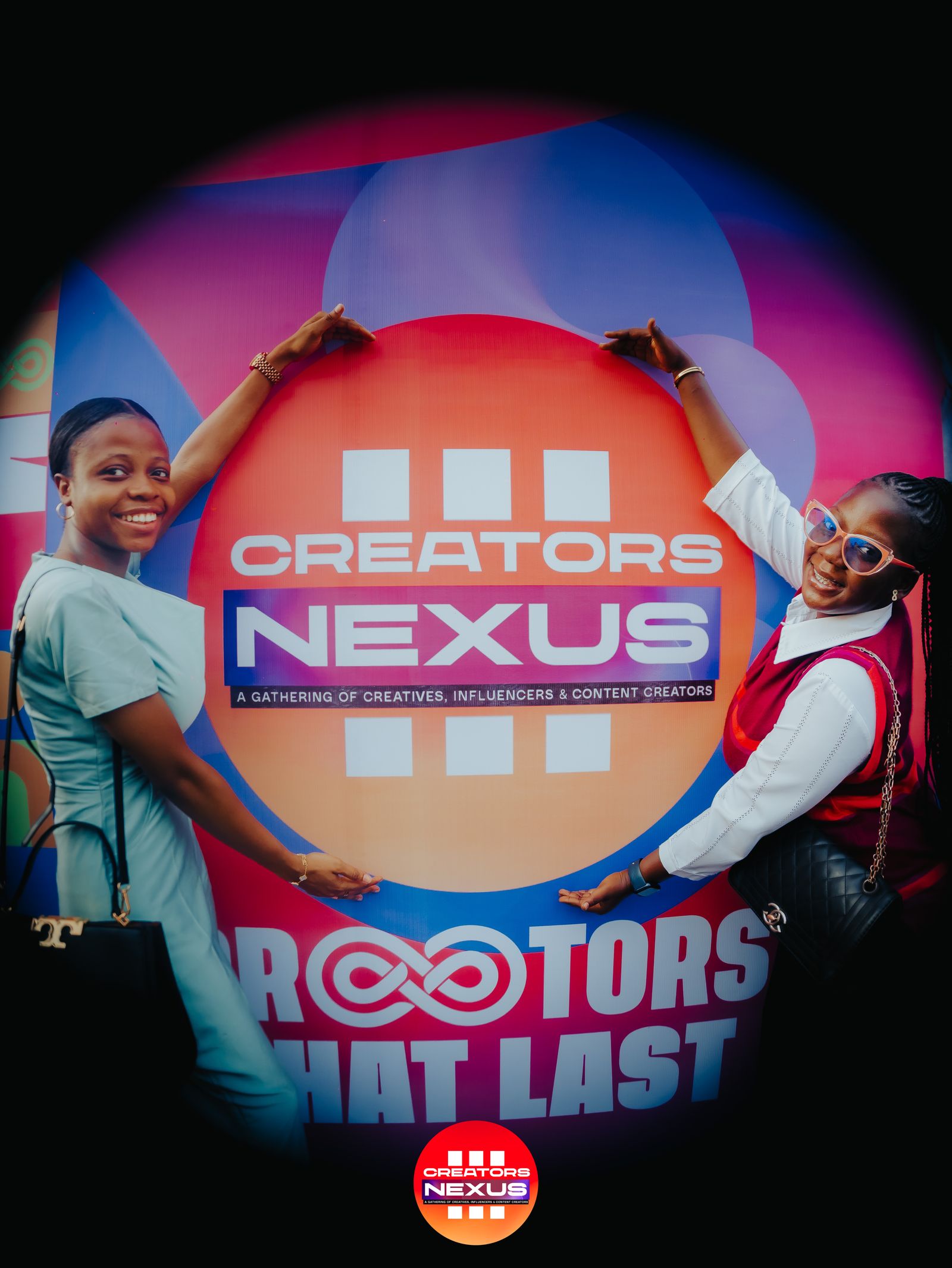 Two attendees posing together in front of the Creators Nexus circular logo backdrop at Creators Nexus III.