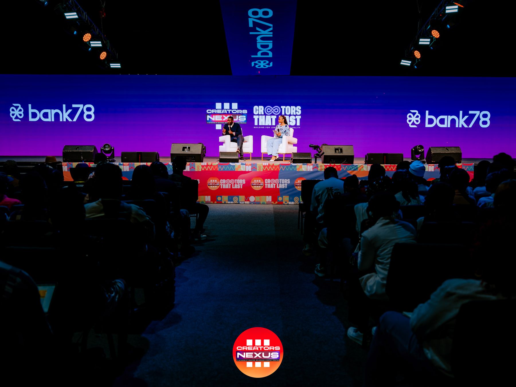 Wide view of the Creators Nexus III main stage during a panel conversation, with Bank78 branding on the screens either side, seen from the audience.